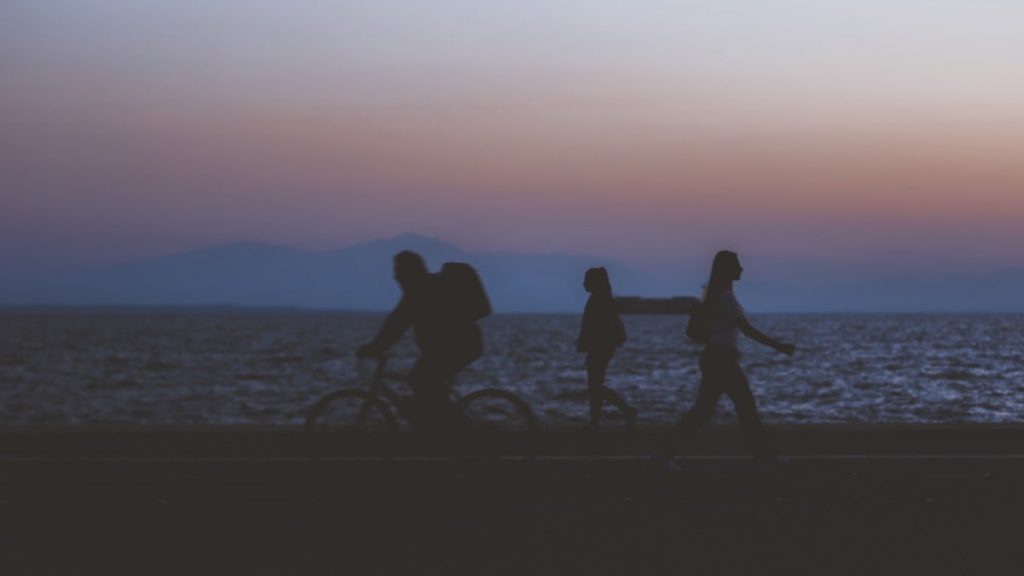 creative unfocused humans wallpaper pattern of fuzzy walking people silhouettes on dark twilight waterfront along Pacific ocean and unfocused mountain horizon background in evening sunset time